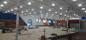 Wide view of The Edge Fitness Clubs construction site showing large open gym area with electrical work underway, including overhead lighting installation, conduit routing, and floor-level power prep performed by our electrical team.