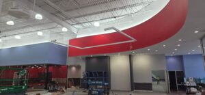 Electrical installation of a suspended rectangular LED light fixture at The Edge Fitness Clubs, showing conduit, ceiling wiring, and the illuminated accent lighting along the curved red wall during construction.