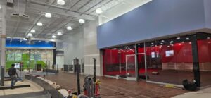 In-progress interior build-out at The Edge Fitness Clubs, showing electrical contractors working around exposed ceilings with newly installed lighting, conduit, and wiring alongside the construction of a red-glass enclosed room.
