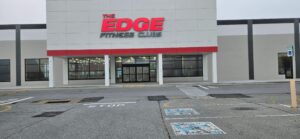 Front exterior of The Edge Fitness Clubs showing finished storefront lighting, new electrical work supporting signage illumination, and upgraded service infrastructure visible through the entrance windows.