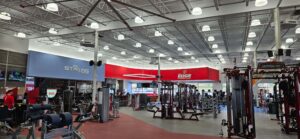 A wide view of The Edge Fitness Clubs gym floor showing extensive overhead electrical work, LED lighting, mounted speakers, and power-fed fitness equipment installed by our electrical team.