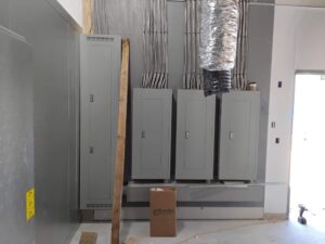 Electrical room inside a Dunkin’ store showing multiple panelboards mounted in a row with neatly routed conduit feeding down from the ceiling, alongside HVAC ductwork and in-progress interior construction.