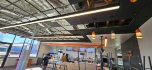 Interior of a Dunkin’ store under renovation showing exposed ceiling grid, installed LED linear fixtures, orange pendant lights, and electrical rough-in work above the front counter area.