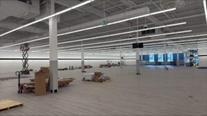 Large retail interior with rows of suspended LED fixtures, new flooring, scissor lifts, and materials staged throughout the space during the final phase of a commercial build-out.