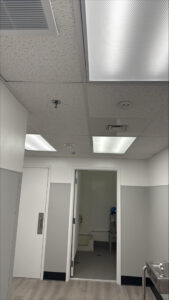 Finished commercial interior showing installed ceiling grid, LED panel lights, HVAC vents, fire alarm devices, and sprinkler heads above a corridor leading into a utility room.