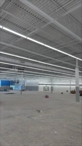 Wide view of a large retail interior with rows of suspended LED fixtures and exposed structural ceiling, showing completed overhead lighting work before flooring and shelving installation.