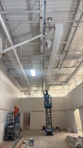Electricians working from scissor lifts install overhead conduit and cable trays in a high-ceiling retail space, with exposed ductwork, lighting, and structural steel visible above.
