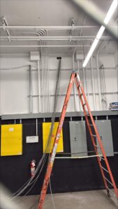 Electrical room with multiple conduits routed across the ceiling and down the wall into panelboards, with an A-frame ladder and loose wiring visible during ongoing installation.