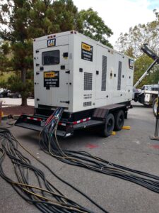 Large CAT rental generator on a trailer with multiple heavy electrical cables routed across the ground, showing a temporary power distribution setup at an outdoor job site.