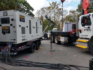 Large CAT rental generator connected with multiple heavy-gauge cables alongside a PSE&G utility truck with a crane boom, showing a temporary power setup in progress at an outdoor job site.
