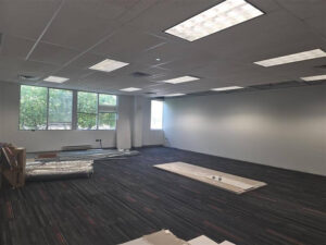 Open office space with installed grid lighting, large windows providing natural light, and construction materials staged on the floor during ongoing renovation work.