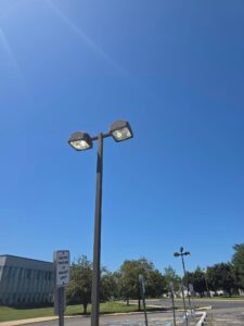 Tall outdoor parking lot light pole with dual LED fixtures illuminated against a clear blue sky, with visitor parking signs and surrounding trees visible in the background.