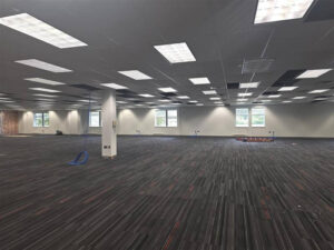 Large open office space with ceiling tiles removed, new lighting installed, and wiring drops and wall outlets in place during early electrical rough-in.