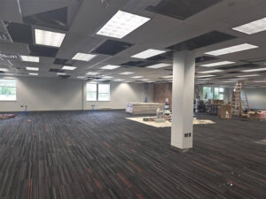 Open office space under renovation with ceiling tiles removed, new grid lighting partially installed, and construction materials and ladders staged throughout the room.
