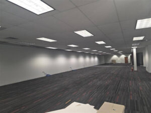 Large open commercial space with new drop-ceiling lighting installed and exposed wall outlets along the perimeter, showing early-stage electrical work during an office build-out.