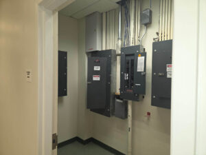 Electrical room viewed from the doorway, showing multiple panelboards mounted on the wall with neatly organized conduit runs and labeling throughout.